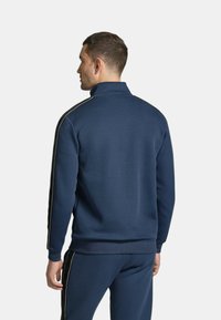 Giacca zip-up blu navy con colletto alto e strisce laterali nere. Texture liscia, polsini e vita a coste, design aderente.