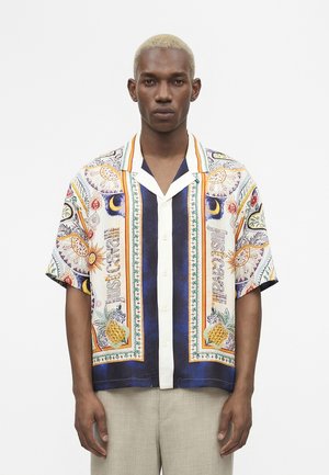 Mladý muž s krátkymi blond vlasmi, ktorý má na sebe farebnú košeľu s krátkym rukávom s gombíkmi a vzormi slnka, mesiaca a ananásu.