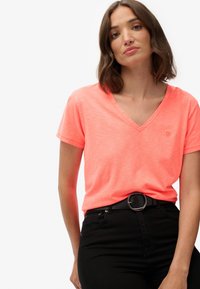 Camiseta de cuello en v color coral neón en tela ligera con manga corta. Lleva un pequeño logo en el pecho. Combinada con jeans negros de cintura alta.