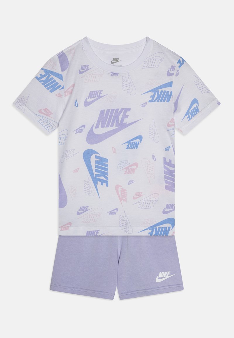Tricou alb din bumbac cu logo-uri Nike multicolore de dimensiuni variate, asortat cu pantaloni scurți violet deschis, prezentând un mic logo Nike.