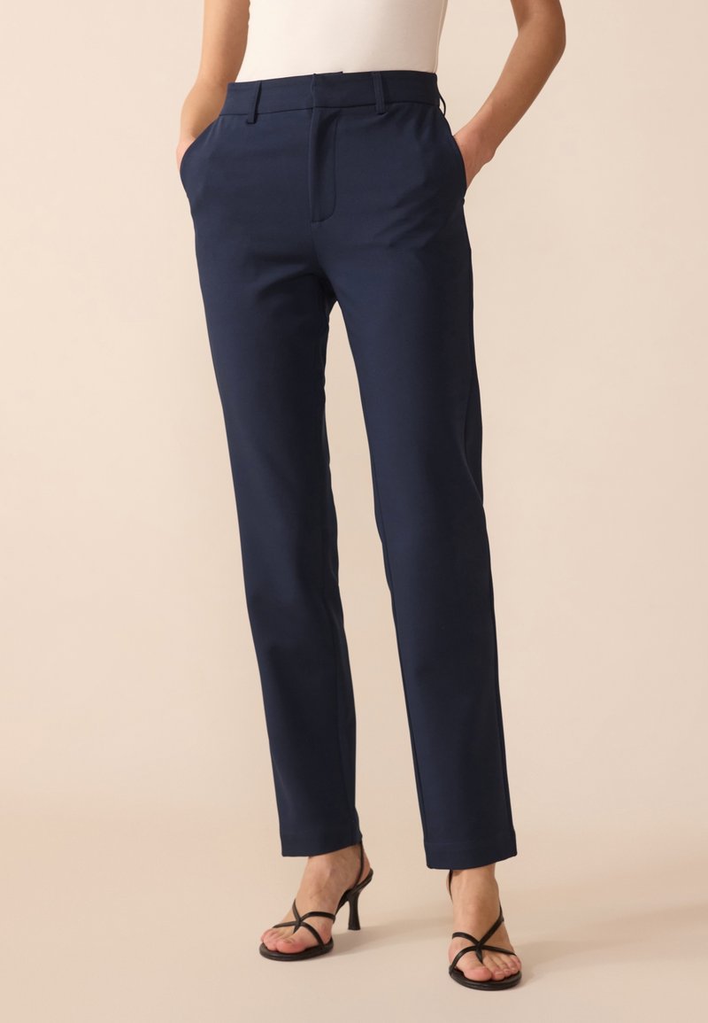 Femme portant un pantalon droit bleu marine avec poches latérales et des sandales à talons ouvertes noires, debout sur un sol beige.