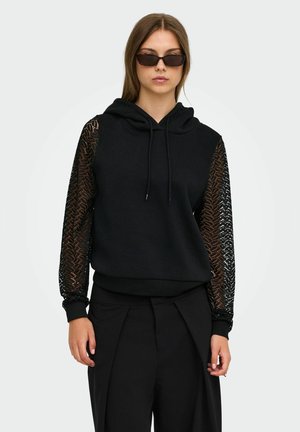 Femme portant un sweat à capuche noir avec des manches en dentelle, un pantalon noir et des lunettes de soleil foncées, debout devant un fond clair.