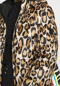 Chaqueta de pluma con estampado de leopardo en un patrón negro y dorado, textura brillante y capucha. Presenta un parche con el logo negro en la manga.