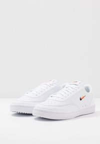 Adidași albi cu partea superioară din piele texturată, vârf perforat și logo Swoosh negru. Prezintă o formă curbată și o talpă din cauciuc cu model de profil.