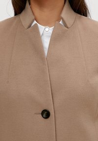 Beige uldfrakke med høj krave, med en enkelt sort knap og minimal syning. Underneath ses en hvid skjorte med krave.