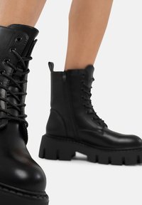 Stivali ankle in pelle nera con suola spessa, chiusura con lacci davanti, zip laterale e superficie texture, caratterizzati da cuciture e occhielli prominenti.