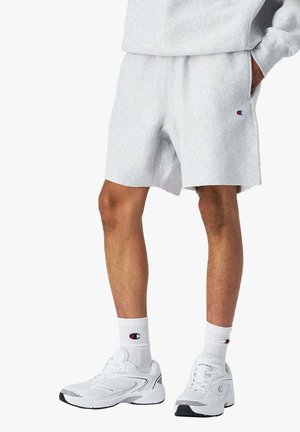 Uomo che indossa pantaloncini Champion grigio chiaro, calzini Champion bianchi e scarpe sportive bianche, in piedi con una mano in tasca.