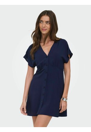 Kvinde med langt brunt hår iført en kortærmet marineblå kjole med knapper foran og sølvfarvet armbånd mod en ensfarvet baggrund.