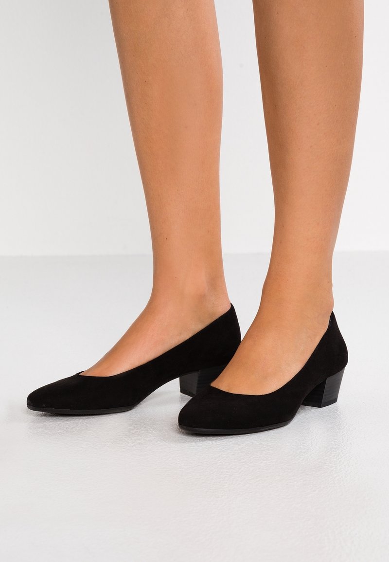 Jambes portant des escarpins à talon bas en daim noir, posées sur un sol clair devant un fond uni.