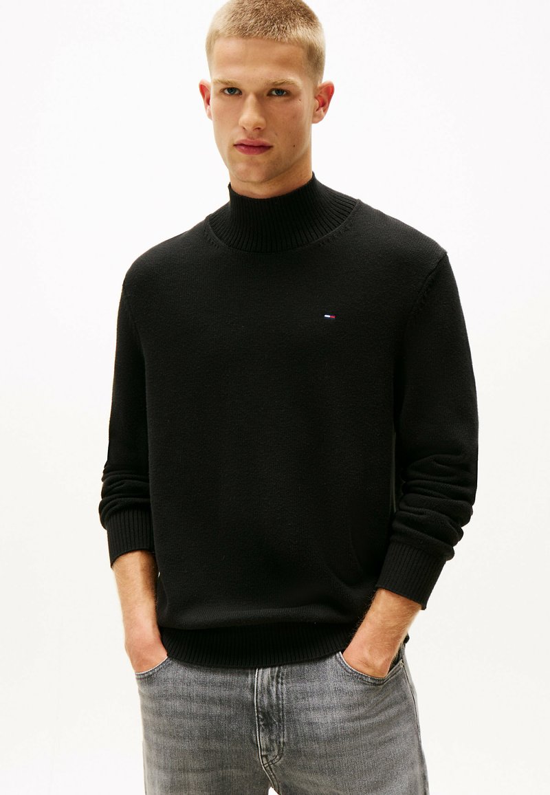 Suéter negro de punto con cuello alto, con puños y dobladillo acanalados, que presenta un pequeño logo en el pecho. El modelo lleva jeans grises y posa con las manos en los bolsillos.