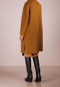 Mosterdkleurige wollen jas met een rechte snit, met een split aan de achterkant, gecombineerd met zwarte leren laarzen tot aan de knie en een tankleurige rok eronder.