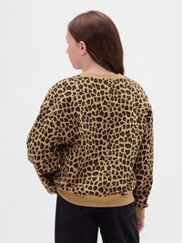 Persona giovane con capelli lunghi e castani, indossa un maglione a stampa leopardata marrone e nera e pantaloni neri, di spalle davanti a uno sfondo bianco.