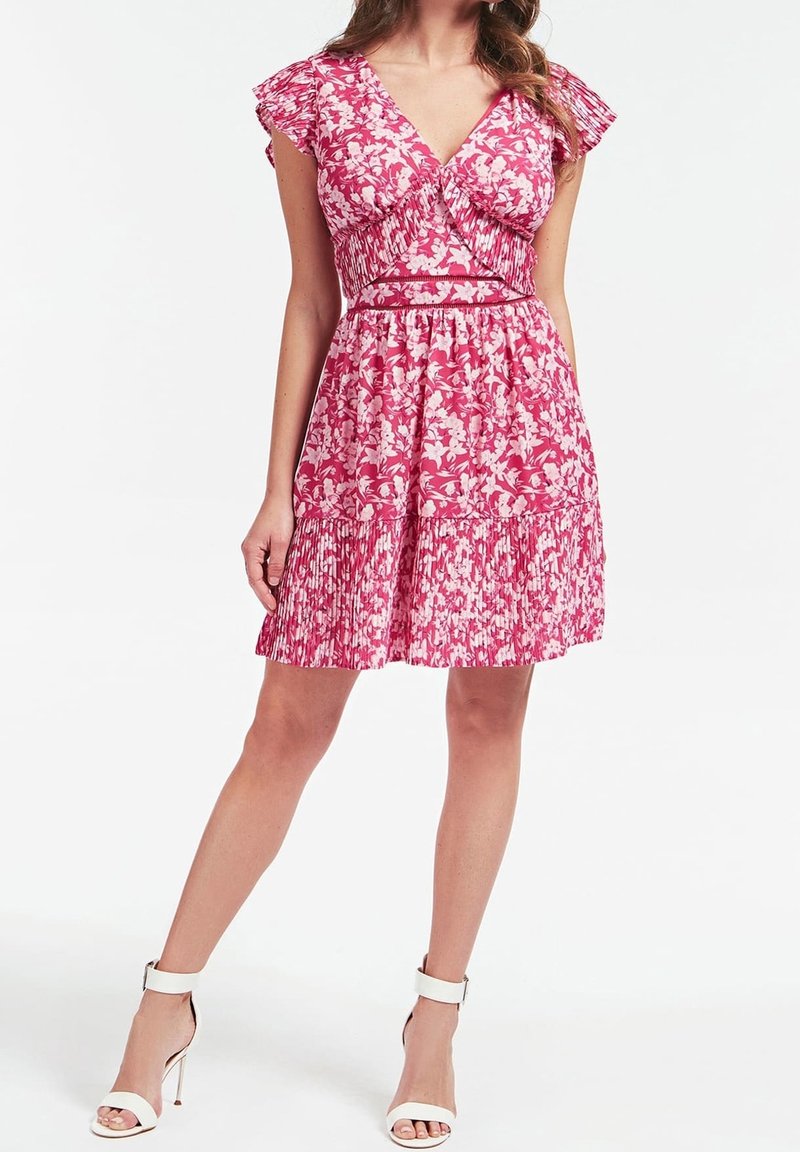 Femme portant une robe courte fleurie rose et blanche avec des manches à volants et un ourlet plissé, associée à des sandales à talons hauts blanches, debout.