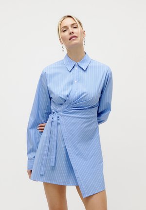 Frau trägt ein hellblau gestreiftes Wickelkleid mit langen Ärmeln und Kragen, steht vor einem schlichten weißen Hintergrund.