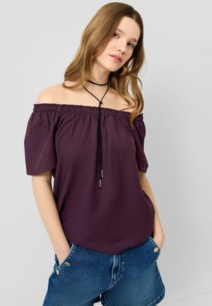 Femme portant un haut violet décolleté aux épaules et un short en jean bleu, avec de longs cheveux, un collier ras du cou noir et les mains dans les poches.