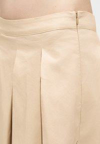 Μπεζ φούστα μέχρι το γόνατο με πλαϊνό φερμουάρ και πιέτες στο μπροστινό μέρος, φορεμένη στη μέση που δείχνει ομαλή υφή υφάσματος.