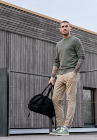 Homme tatoué portant un pull vert et un pantalon beige, tenant un sac de sport noir, debout sur une terrasse en bois devant un bâtiment moderne.