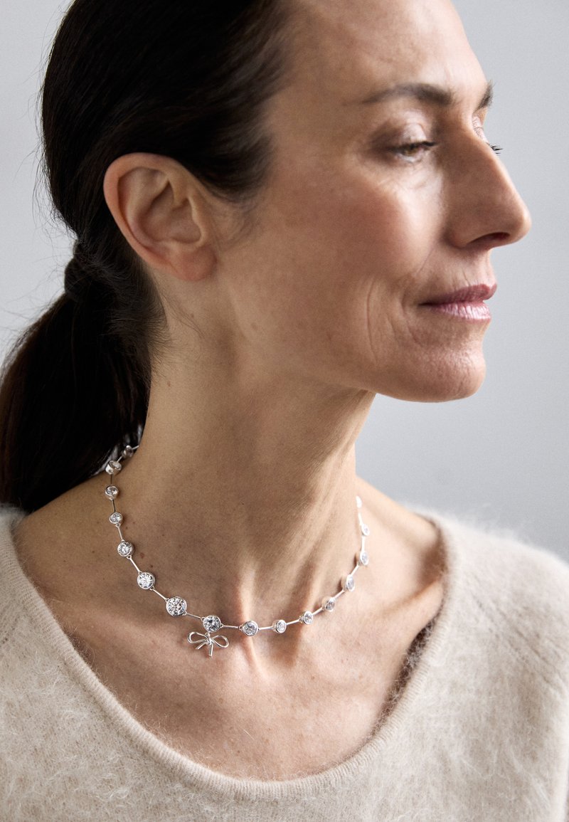 Ασημένιο κολιέ με στρογγυλούς κρύσταλλους και λεπτομέρεια σε σχήμα φιόγκου, με απαλό, μεταλλικό φινίρισμα. Φορεμένο πάνω από ένα ουδέτερο πουλόβερ.