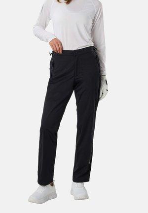 Person iført sorte bukser med lynlås, hvid langærmet skjorte, hvid handske på venstre hånd og hvide sneakers står på en ensfarvet baggrund.