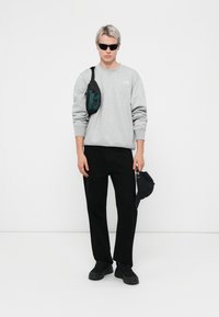 Grijze sweatshirt, zwarte broek en zwarte sneakers. Zwarte schoudertas met een groene accent. Zwarte pet in de rechterhand vastgehouden. Minimalistisch ontwerp.
