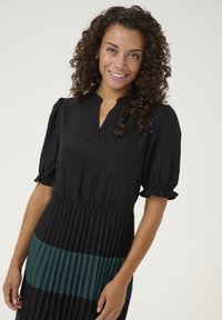 Čierne a tyrkysové plisované šaty s výstrihom do V a krátkymi naberanými rukávmi. Hrudník je priliehavý, prechádzajúci do voľnej, rebrovanej sukne.