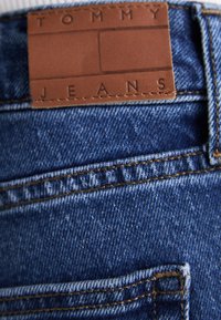 Denim jeans with a light blue wash, featuring prominent stitching, a small back pocket, and a brown leather label embossed with "TOMMY JEANS."