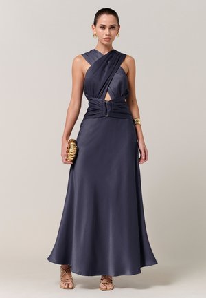 Vrouw draagt een mouwloze donkerblauwe satijnen jurk met een gedraaide banddetail, houdt een goudkleurige textuurtas vast en draagt gouden oorbellen en sandalen.