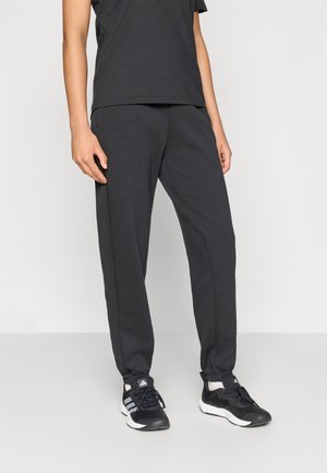 Dolna część ciała osoby ubranej w czarne spodnie dresowe, czarne buty sportowe Adidas z białymi paskami i czarną bluzkę na jednolitym tle.