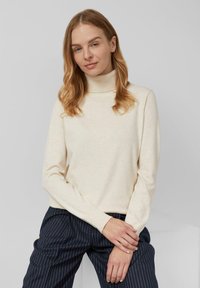 Maglione a collo alto beige realizzato in morbido tessuto a maglia con maniche lunghe, abbinato a pantaloni navy a righe sottili, su uno sfondo neutro.