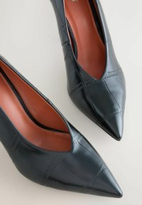 Fekete, hegyes orrú bőr női cipő pár, varrott keresztmintával és barna belső béléssel.