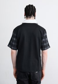 Black short-sleeve polo with a white collar and plaid fabric on the sleeves. Made of smooth material, featuring a relaxed fit and casual design.