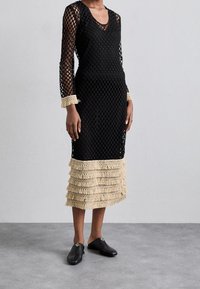 Abito midi nero con maniche lunghe trasparenti, dettagli in frangia crema su polsini e orlo, indossato con scarpe slip-on nere su pavimento grigio.