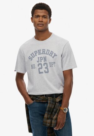 Mężczyzna w jasnoszarej koszulce "Superdry JPN No. 23 DPT", niebieskich jeansach, koszuli w kratę związanej w talii oraz brązowym zegarku na nadgarstku.