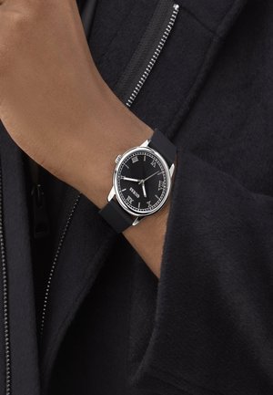 Black analog watch with silver accents, Roman numeral markers, and a black rubber strap, worn on a hand with a zipper coat.