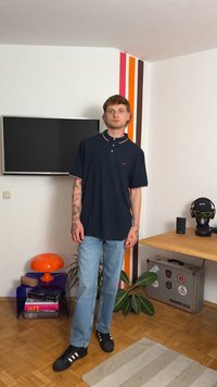 Jūras zila polo krekls ar baltu un sarkanu apkakles apdari, kombinēts ar gaiši ziliem džinsiem un melniem apaviem. Telpa ar televizoru, oranžu lampu un augiem.