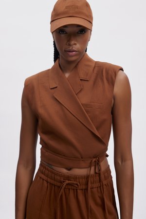 Gilet corto marrone con design doppiopetto e una piccola tasca, abbinato a pantaloni con coulisse coordinati e un berretto marrone con logo.