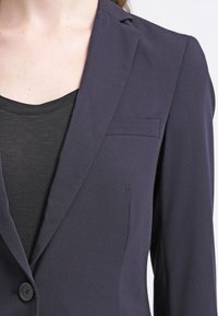 Námorně modrý blejzr s přiléhavým střihem, jednostranným zapínáním na knoflík a malou náprsní kapsou. Tkanina se zdá být hladká s matným povrchem.