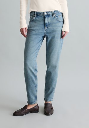 Dolna część ciała osoby ubranej w jasnoniebieskie proste dżinsy, kremową bluzkę z długim rękawem oraz ciemnobrązowe mokasyny stojącej na szarym tle.