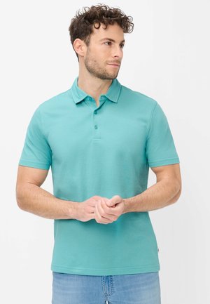 Mann mit lockigem Haar, der ein türkisfarbenes kurzärmeliges Poloshirt und hellblaue Jeans trägt, die Hände zusammenhaltend, schaut zur Seite.