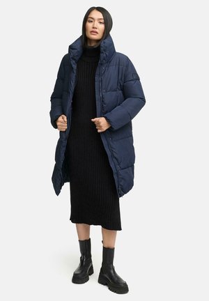 Mörkblå puffjacka med hög krage, med en vertikal quiltad design och lång längd, kombinerad med en svart ribbad klänning och svarta klumpiga stövlar.