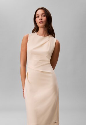 Nő hosszú barna hajjal, ujjatlan krémszínű ruhát visel, és egy homogén világosszürke háttér előtt áll.