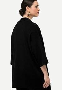 Př oversized černý svetr s kulatým výstřihem, krátkými rukávy a měkkou texturou. Zadní pohled ukazuje uvolněný střih a minimalistický design.