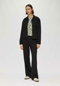 Zwarte cropped jack met een knoopsluiting en twee borstzakken over een geprint top. Zwarte wijde broek en beige sneakers maken de look compleet.