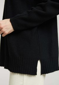 Pull en maille noir avec des poignets côtelés et des fentes sur les côtés, fabriqué à partir d'un matériau doux, avec une coupe décontractée et une texture lisse.