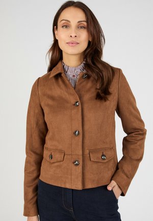 Femme portant une veste marron à boutons par-dessus un haut rayé à col montant, avec une main dans la poche d'un jean bleu foncé, faisant face à l'avant.