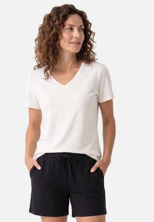 Frau mit lockigem Haar, die ein weißes kurzärmeliges V-Ausschnitt-Shirt und schwarze Shorts mit Kordelzug trägt, Hände in den Taschen, schaut nach rechts.
