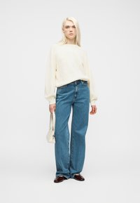 Νεαρή γυναίκα στέκεται, φορόνττας εκρού πουλόβερ με πλεξούδες, άνετο μπλε τζιν, μπορντό παπούτσια και κρατώντας μια μικρή λευκή τσάντα.