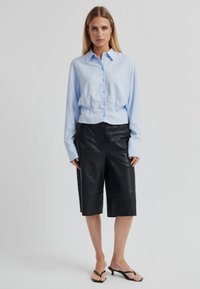 Světlomodrá košile na knoflíky s dlouhými rukávy a zkráceným střihem, doplněná černými koženými krátkými kalhotami po kolena a černými žabkami.