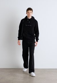 Zwarte pullover hoodie met een logo, gecombineerd met zwarte broek en witte sneakers. Het model staat tegen een effen achtergrond.