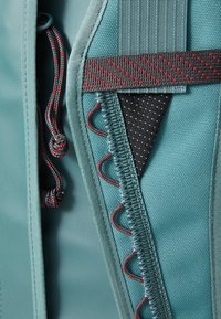 Teal fabric with textured accents. Features red and black stitching, elastic cord, and a patterned inner pocket. Versatile, durable materials used.